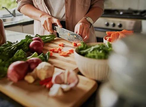 Person chopping orange vegetables on a wooden cutting board in a kitchen surrounded by fresh ingredients including garlic, radishes, green leafy vegetables, and herbs.