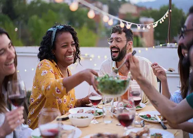 A group of friends laughing and sharing a salad at an outdoor dinner table with lit string lights and wine glasses.