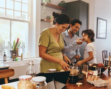 A family in a kitchen with a pregnant woman preparing food, a man feeding a young child, and various kitchen items on the counter including eggs and vegetables.