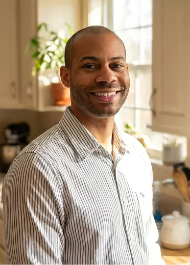 Smiling man with a bald head and trimmed beard wearing a white and black striped shirt standing in a sunlit kitchen with potted plants and kitchenware in the background.