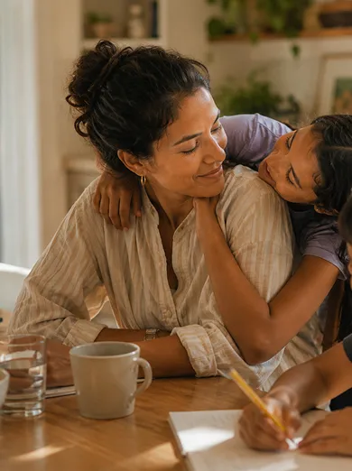 A mother smiling warmly at her daughter who hugs her from behind, while a boy writes with a pencil at a table in a cozy kitchen setting.