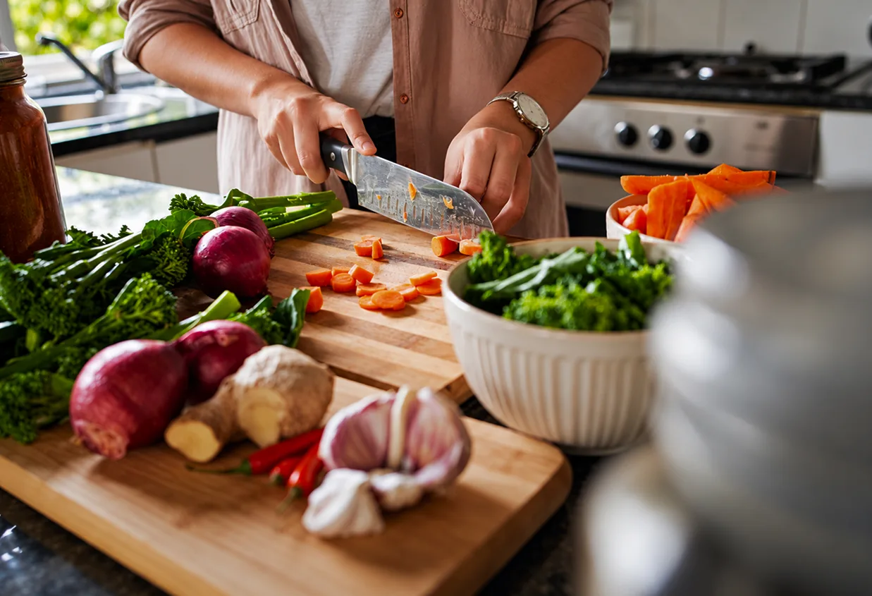A person slicing carrots on a wooden cutting board in a home kitchen, surrounded by red onions, broccolini, ginger, garlic, chili peppers and a bowl of greens.
