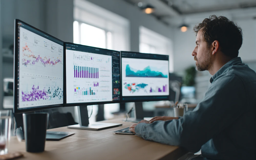 Professional data analyst working at a modern desk with three monitors displaying data visualizations, graphs, and charts during the data cleansing process to ensure accurate and reliable business intelligence.