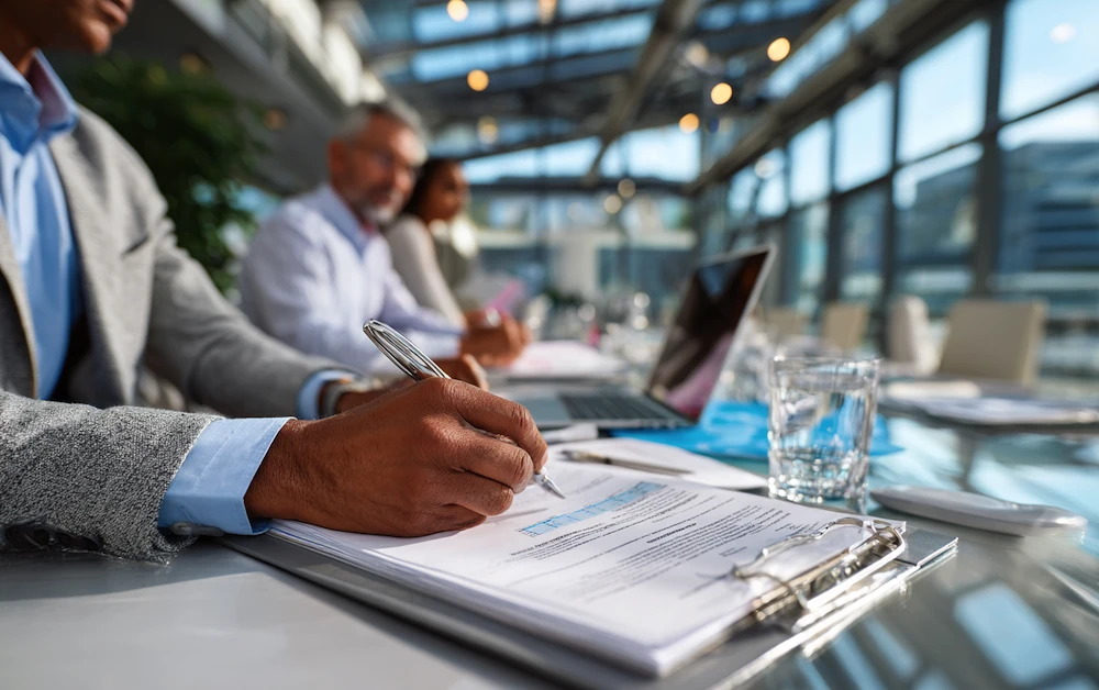 Business professionals conducting an integrated audit review at a modern glass-walled office, analyzing documents with financial data, laptops, and reports on a boardroom table.