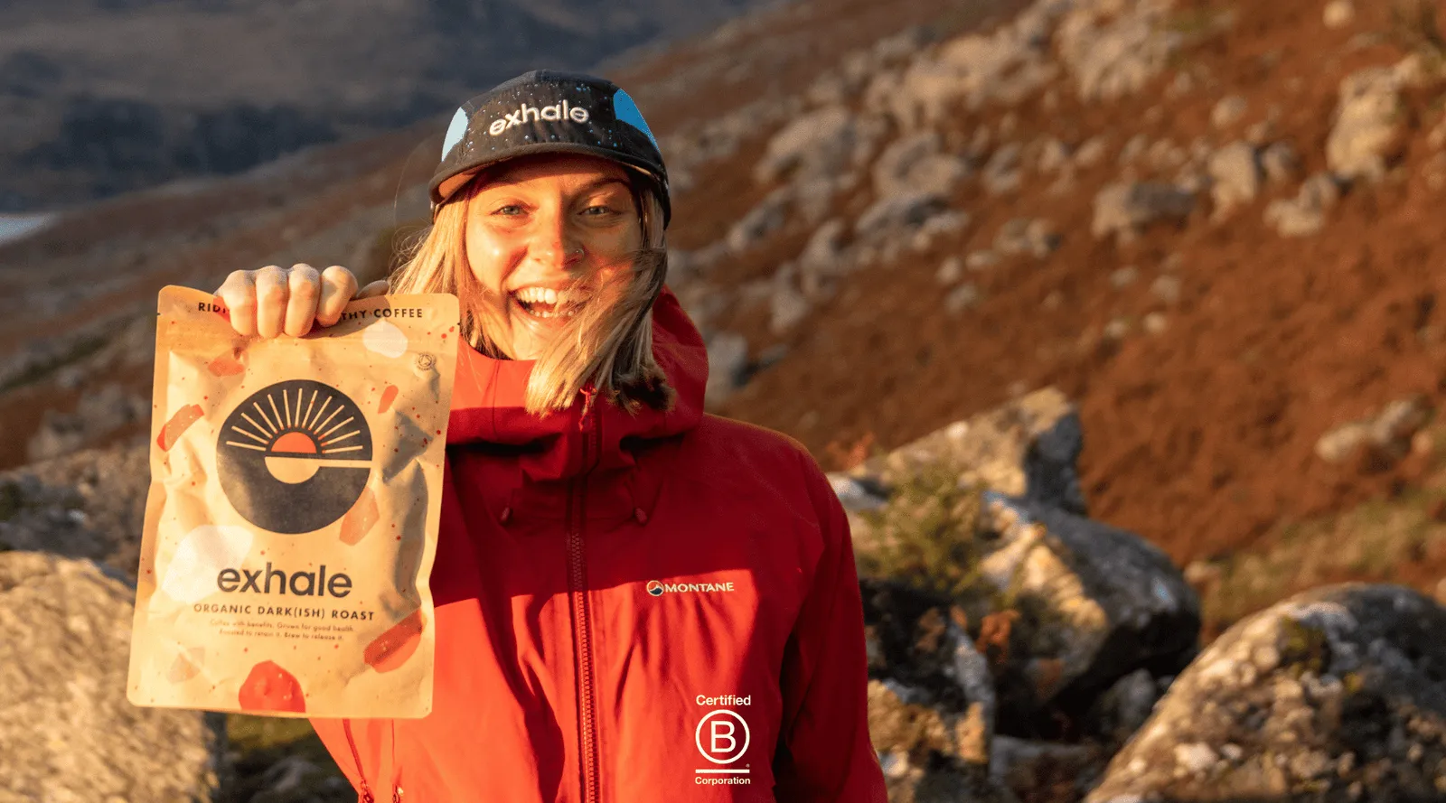 A smiling hiker on a sunlit hillside holding up a bag of Exhale Organic Dark(ish) Roast coffee