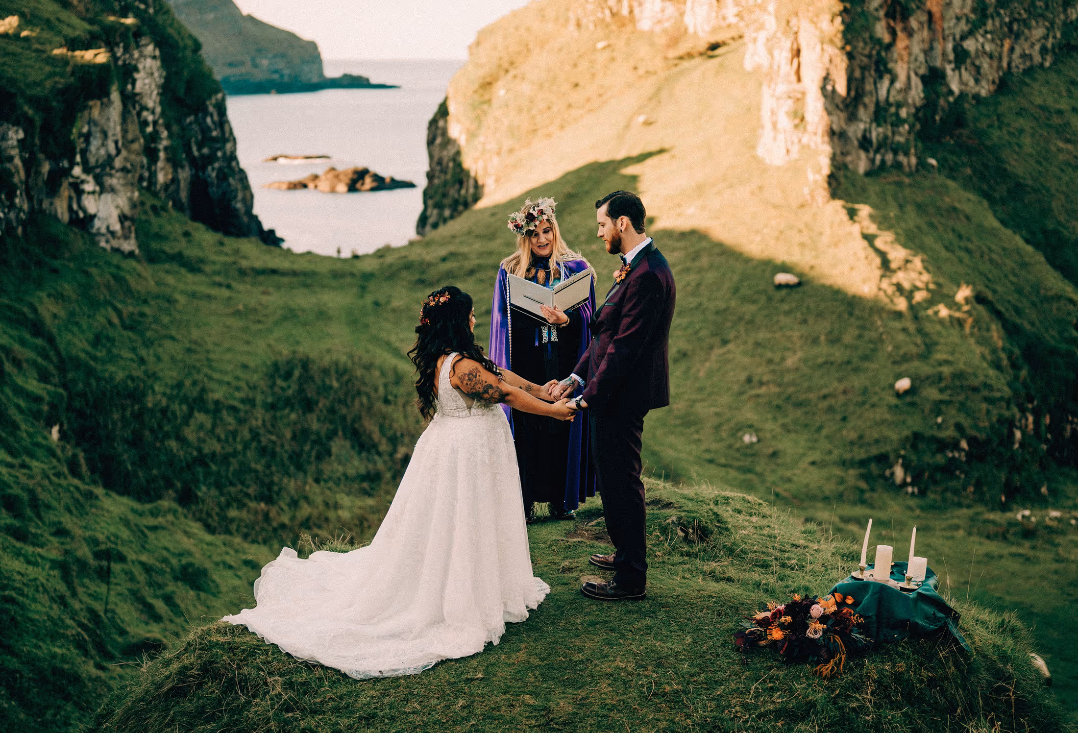 Bride and groom holding hands during an outdoor wedding ceremony on grassy cliffs with an officiant reading from a book.