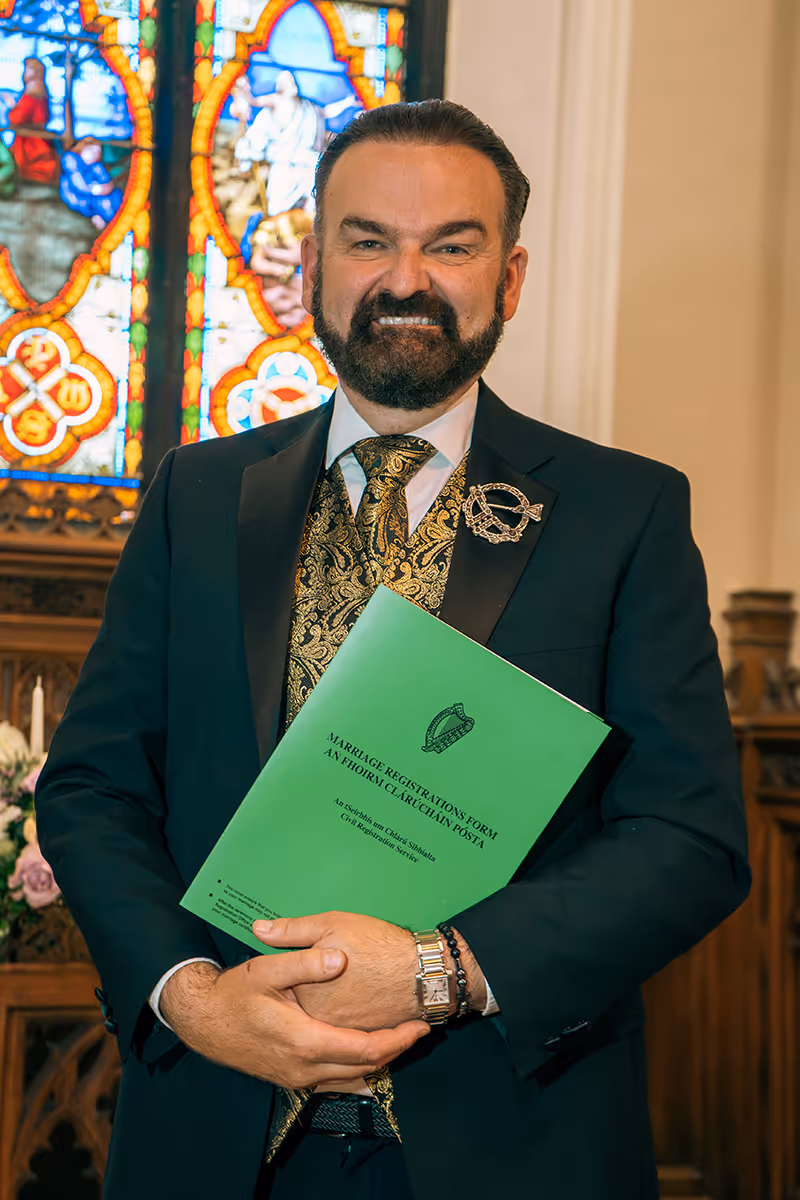 Smiling man in formal suit holding a green Marriage Registrations Form inside a church with stained glass windows in the background.