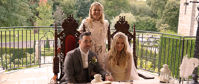 Bride in white dress and veil and groom in gray suit seated in ornate wooden chairs with a smiling woman standing behind them on a terrace with greenery in the background.
