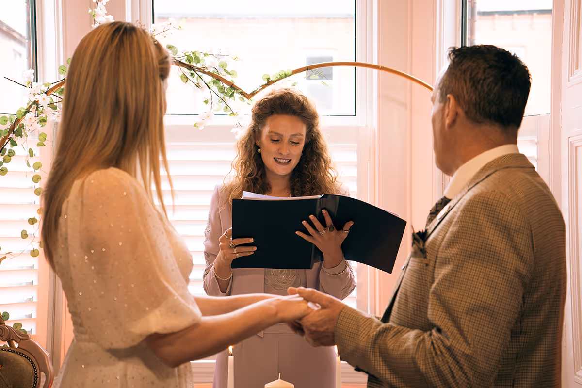 A couple holding hands facing an officiant who is reading from a binder during a wedding ceremony by a window with floral decor.