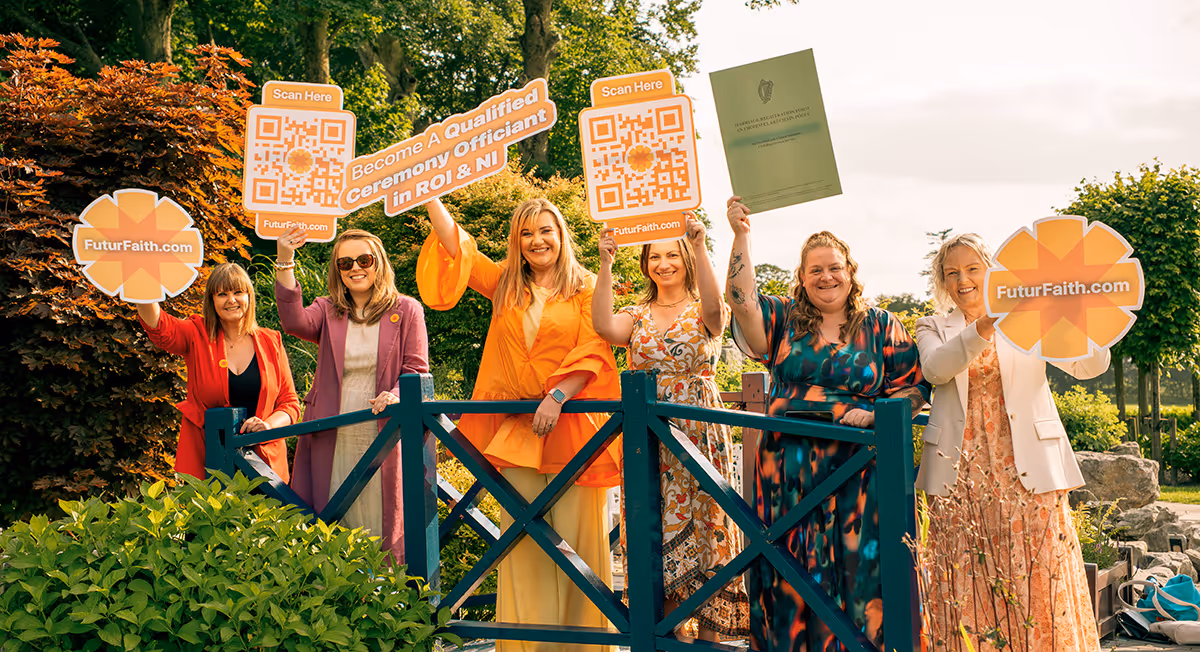 Six women standing behind a blue wooden fence outdoors, holding signs promoting FuturFaith.com and certification as ceremony officiants in ROI and NI.
