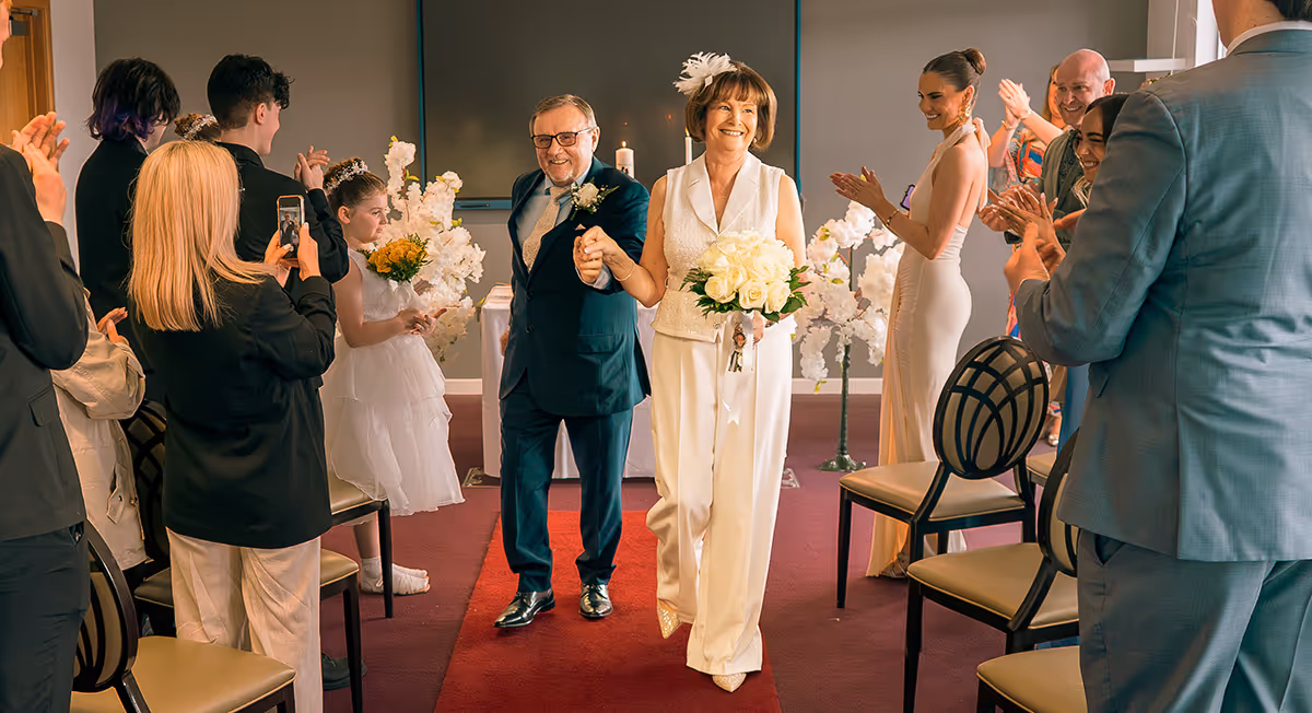 A smiling older couple holding hands and walking down an aisle with guests clapping and a flower girl holding a bouquet nearby.