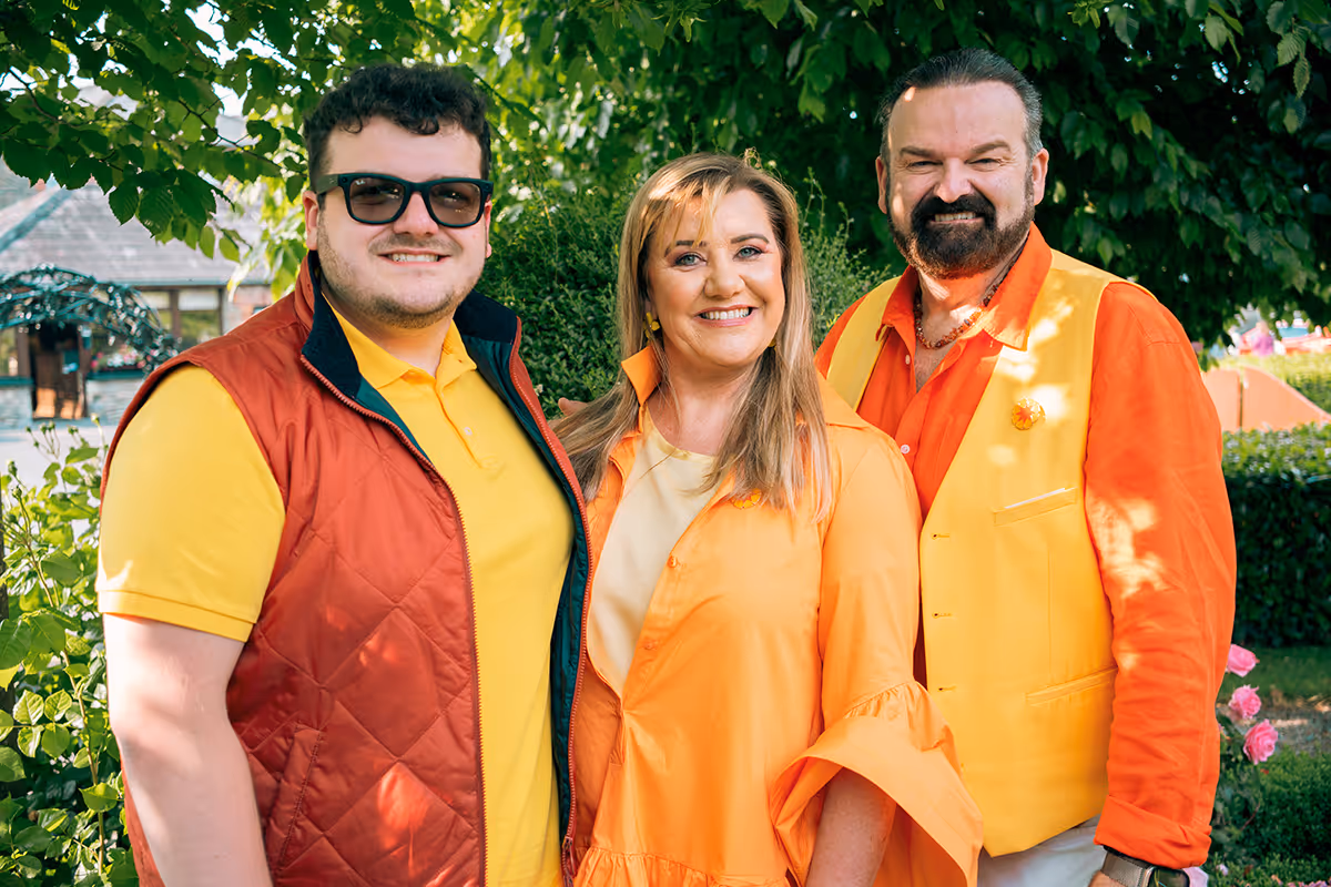 Three smiling people standing outdoors in bright yellow and orange clothing with green foliage in the background.