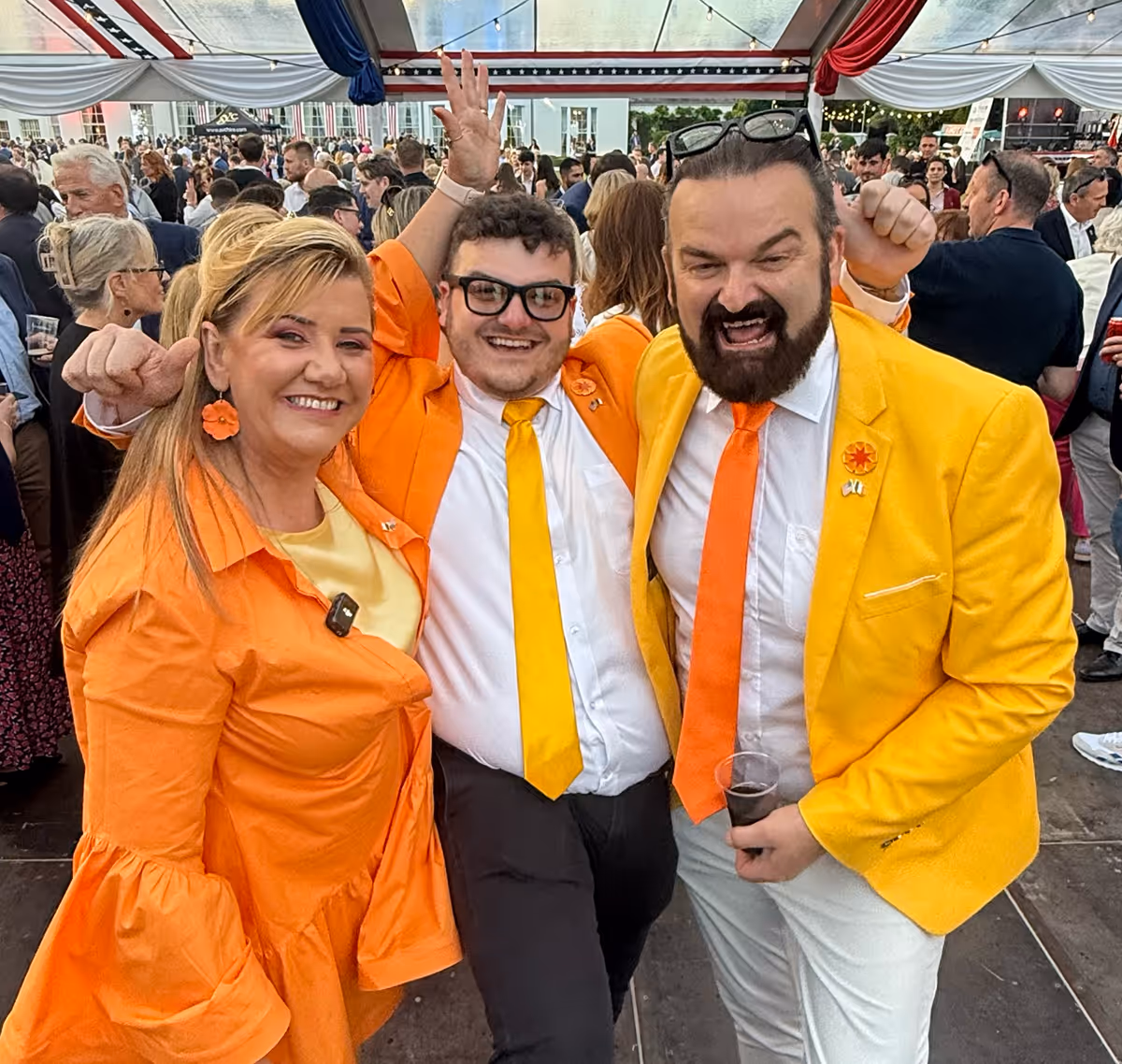 Three adults dressed in bright orange and yellow clothing smiling and posing energetically at a crowded event under a tent.