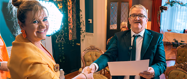 Woman in yellow jacket shaking hands with a man in a suit holding a certificate in a warmly lit room.