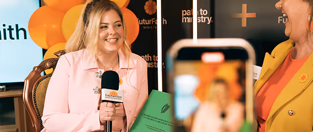 Smiling woman in a pink blazer holding a microphone and green document during an interview setting.