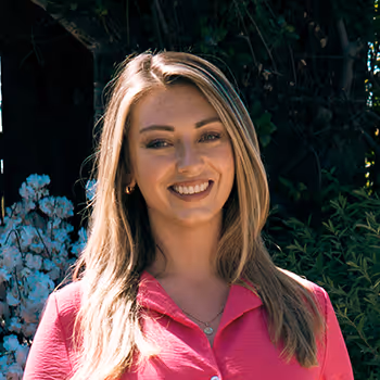 Woman with long blonde hair smiling, wearing a pink shirt, standing outdoors with greenery in the background.