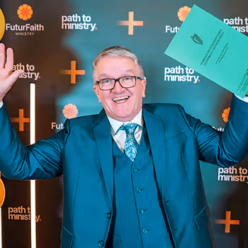 Smiling man in teal suit and glasses holding up a green document, standing in front of a backdrop with 'FuturFaith Ministry' and 'path to ministry' logos.