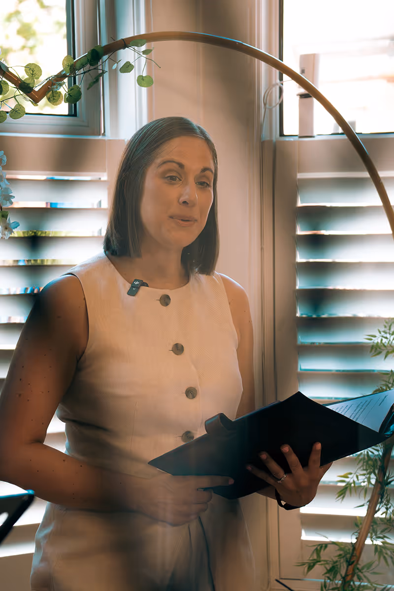 Woman in a sleeveless beige dress holding a folder or book, standing indoors near windows with white blinds and some greenery.