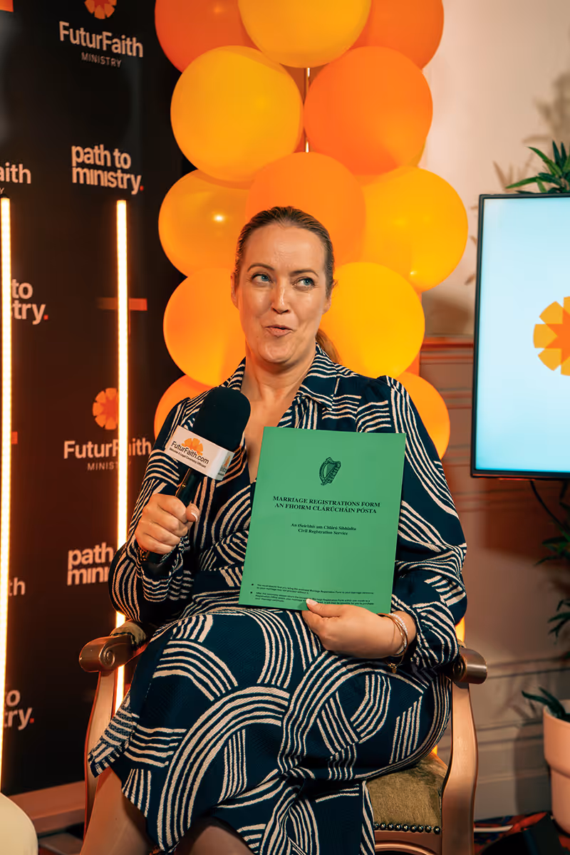 Woman in patterned dress seated holding a green Marriage Registrations Form and a microphone with FuturFaith.com label, with orange balloons and FuturFaith Ministry backdrop behind her.