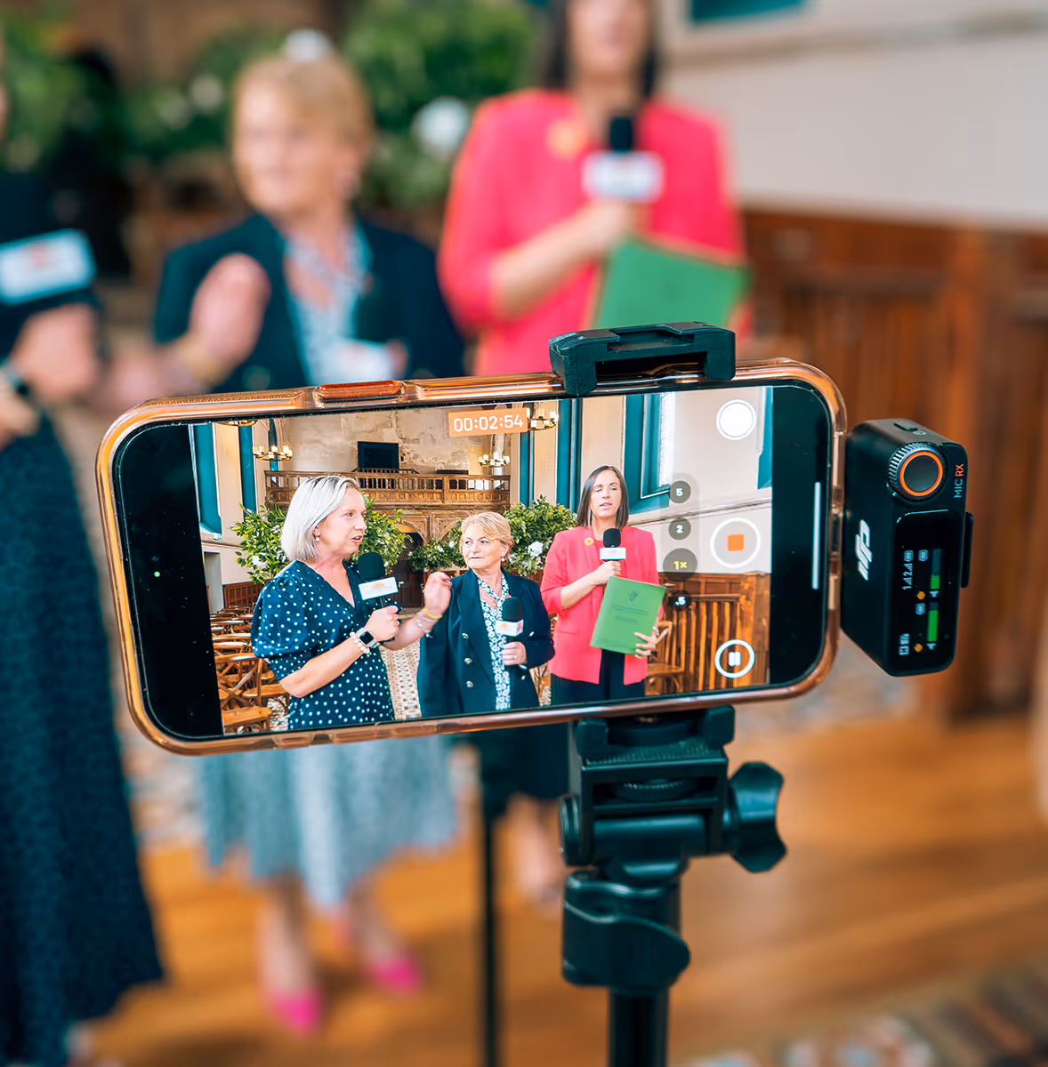 Smartphone on tripod livestreaming three women holding microphones inside a room with wooden chairs and plants.