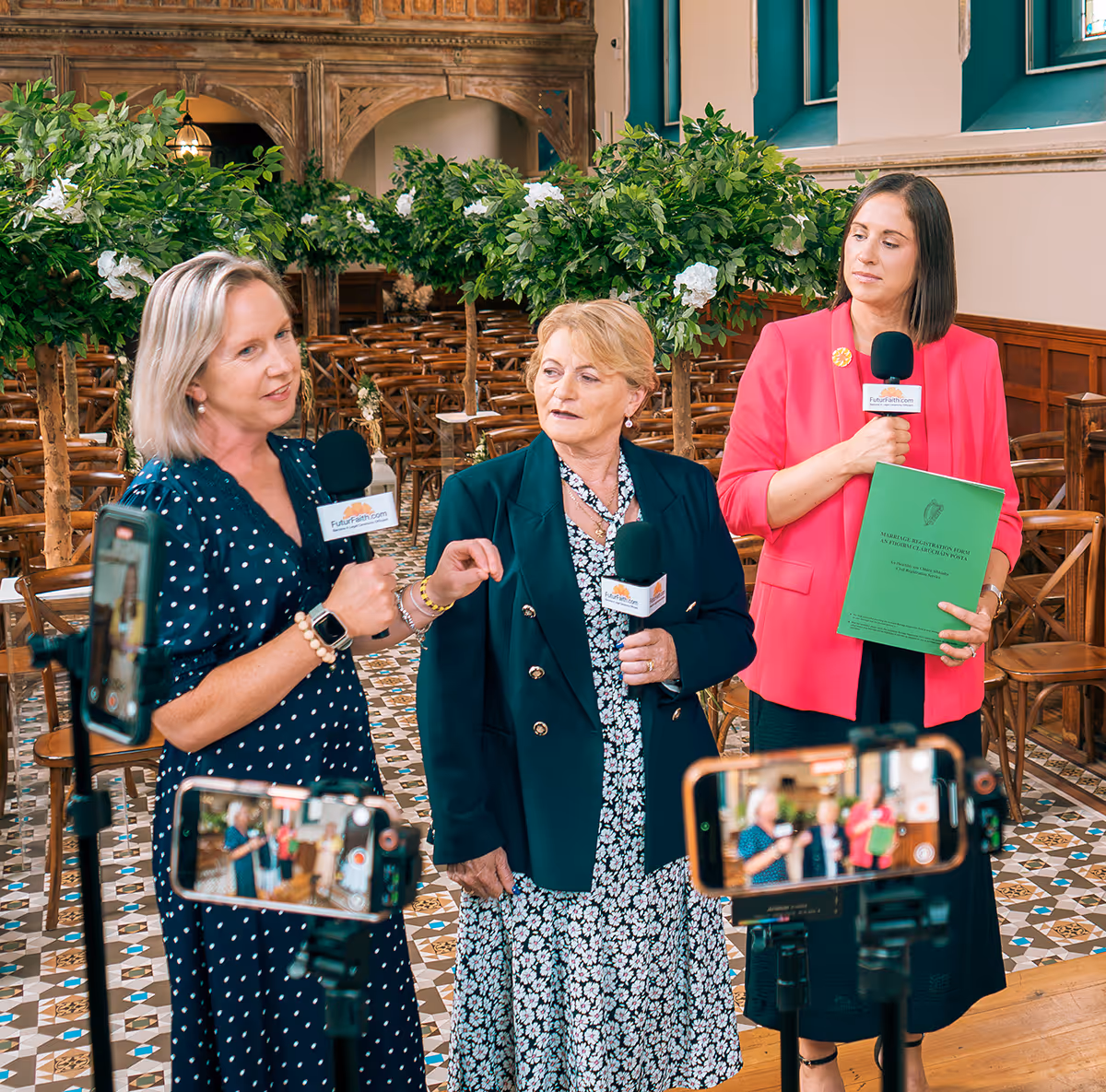 Three women holding microphones and recording a discussion inside a decorated venue with wooden chairs and green plants.
