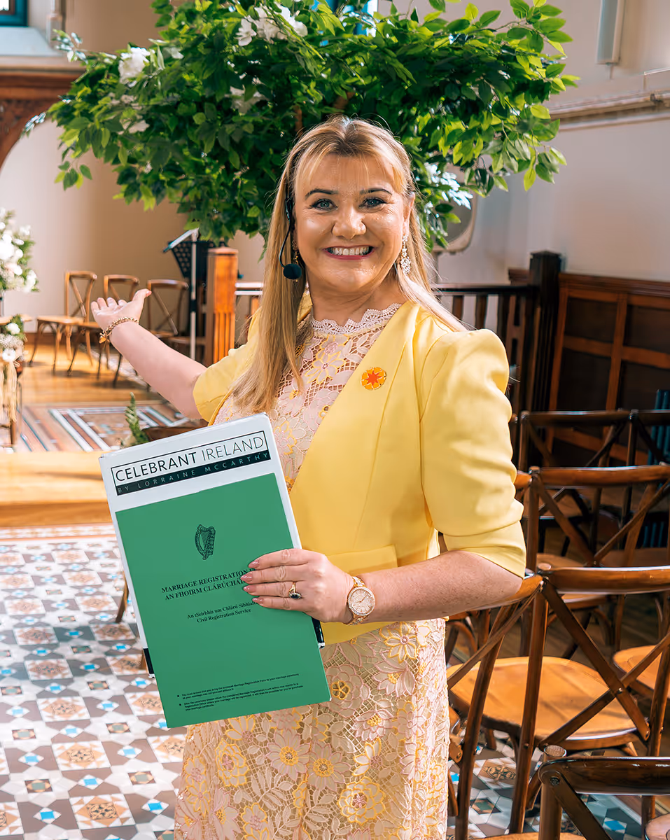 Smiling woman in yellow lace dress and jacket holding a Marriage Registration folder inside a decorated room with wooden chairs.