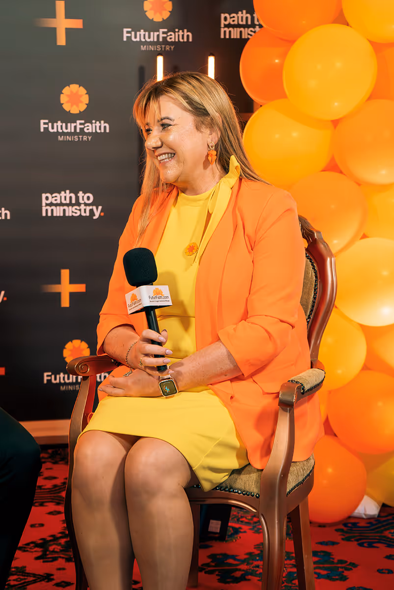 Smiling woman in a yellow dress and orange blazer sitting on a chair, holding a microphone in front of a backdrop with FuturFaith Ministry logos and orange balloons.