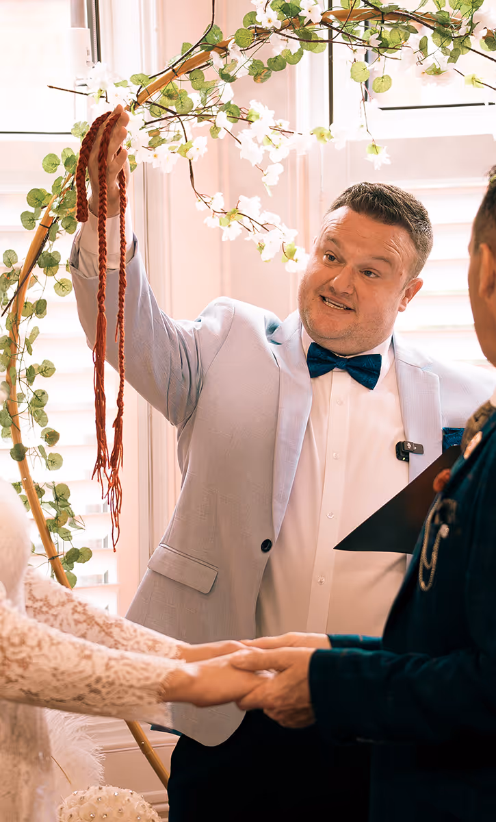 Wedding officiant holding braided cords above a couple holding hands during a ceremony with floral decor in the background.