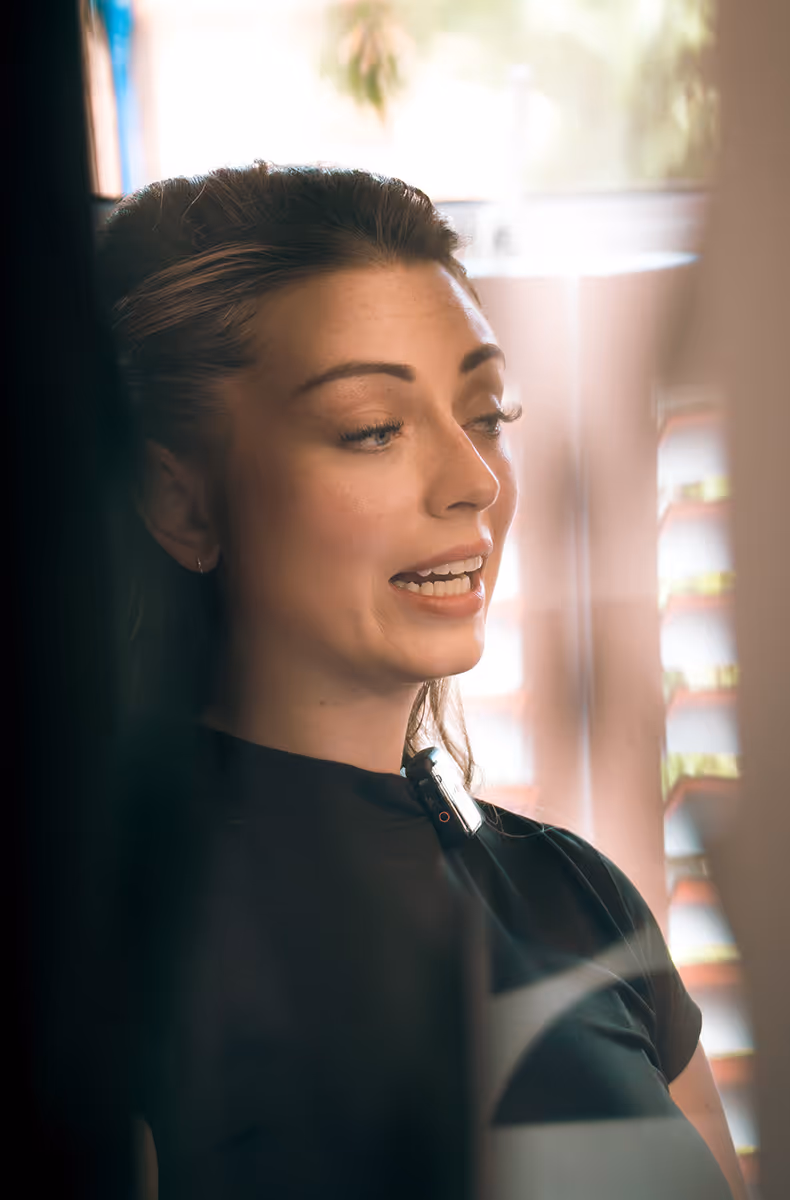 Close-up of a woman speaking, wearing a black top with a microphone clipped to it, in a softly lit indoor setting.