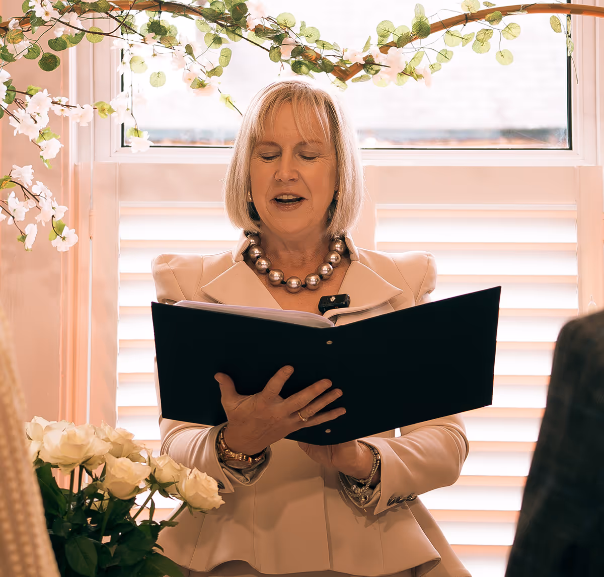 Woman with short blonde hair wearing a pearl necklace and beige blazer reading from a black folder indoors near a window decorated with white flowers.