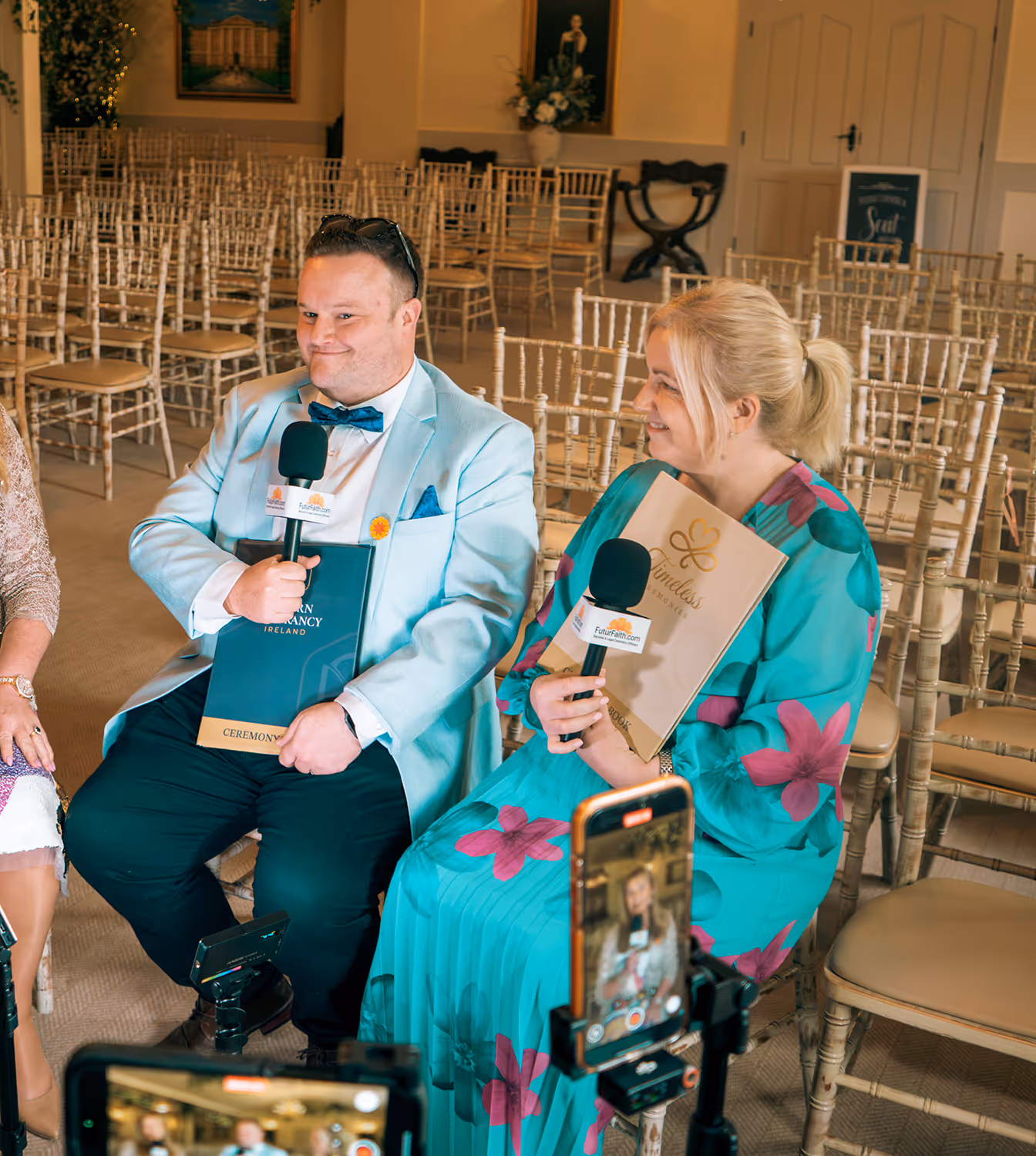 Man in light blue suit and woman in turquoise dress with pink flowers holding microphones and ceremony folders seated in a room with empty chairs and recording phones.