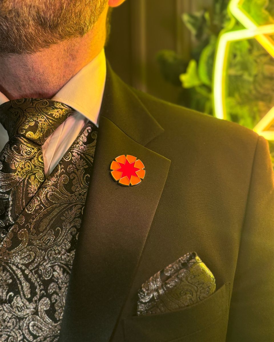 Man wearing black paisley patterned tie and pocket square with a suit jacket adorned by a red and orange enamel lapel pin.