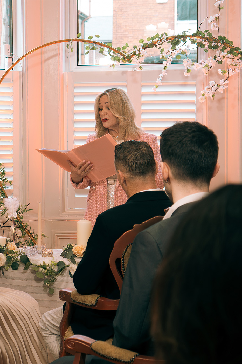 Woman in pink jacket reading from a folder during a formal indoor event with seated attendees.