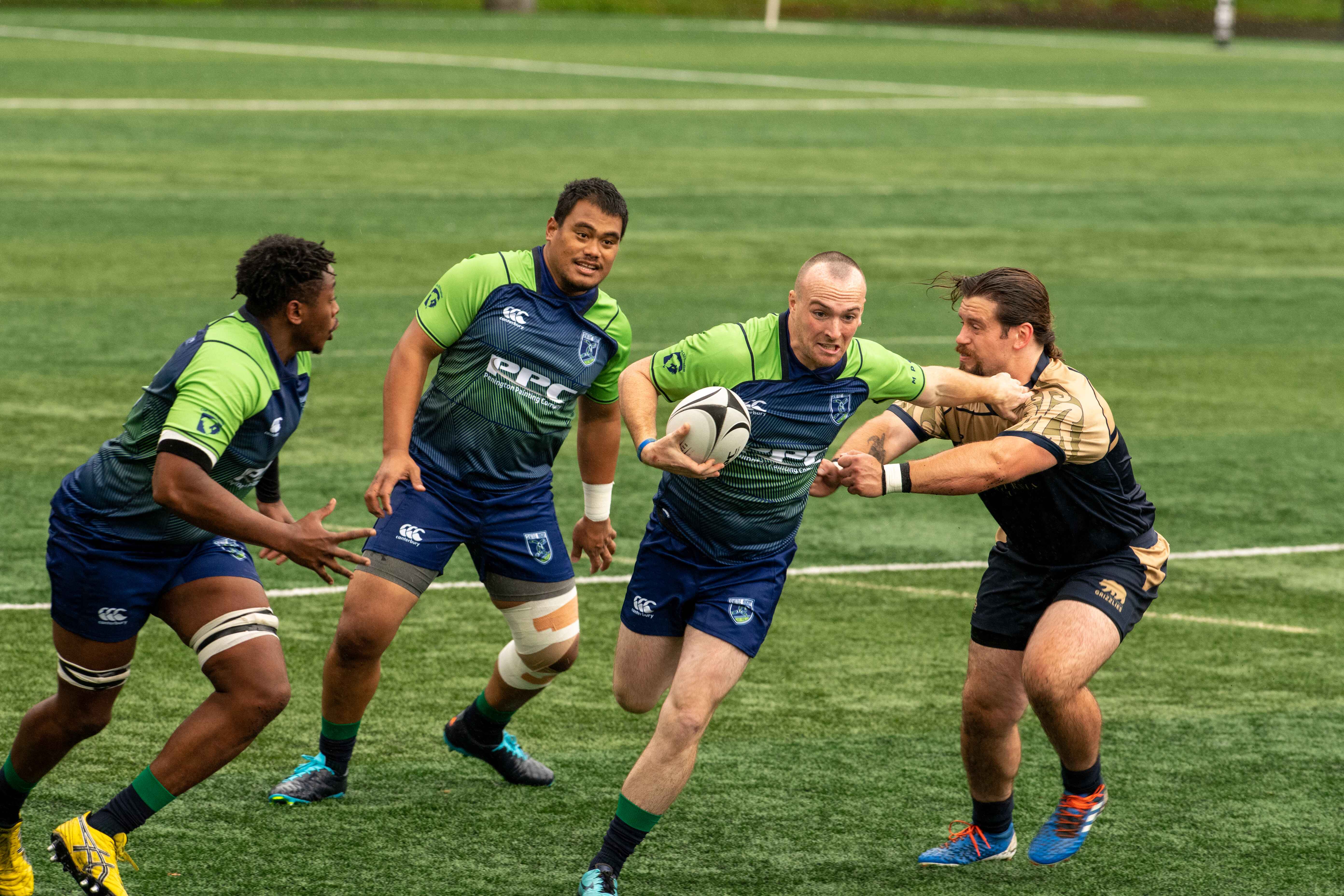 Rugby player in blue and green runs with ball while fending off opponent in black and gold jersey.