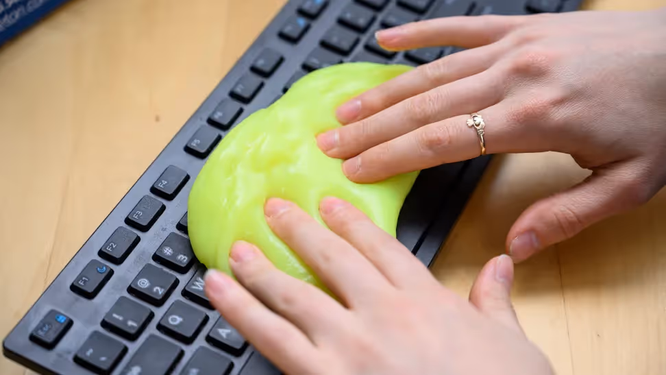 Two hands apply cleaning gel to a black keyboard on a wood desk