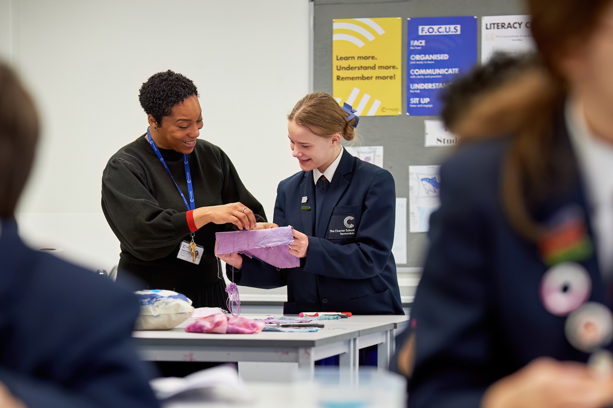 A teacher and a student in a classroom engage in a fabric craft activity, with the teacher guiding the student holding a purple cloth. Both are smiling, and other students and craft materials are partially visible nearby.