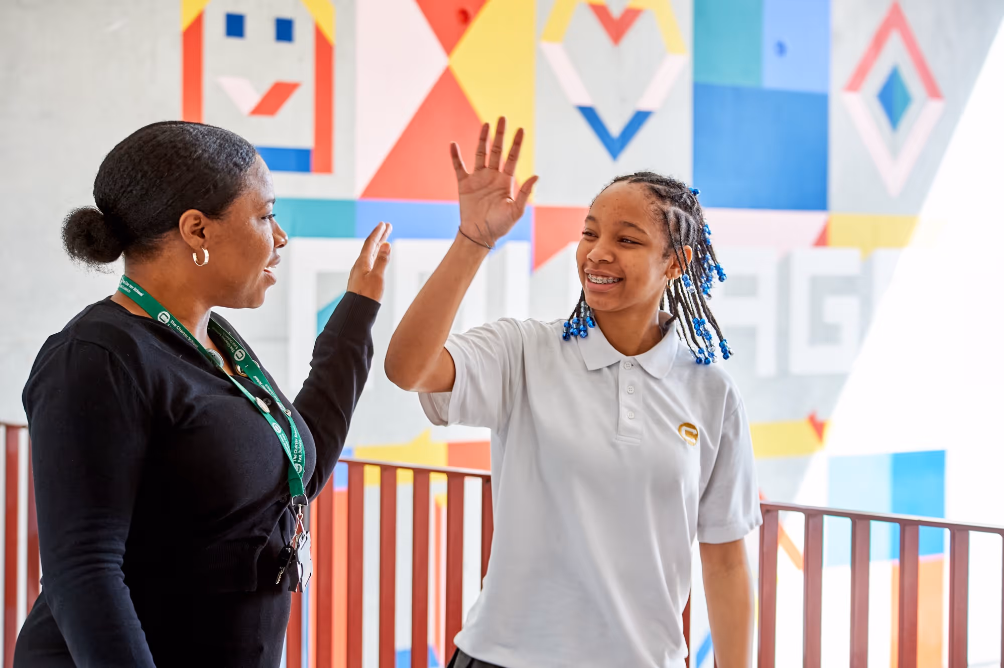 A young girl with braided hair and a white polo shirt gives a high five to a woman with a black long-sleeve shirt and a green lanyard, standing in front of a colorful geometric mural.
