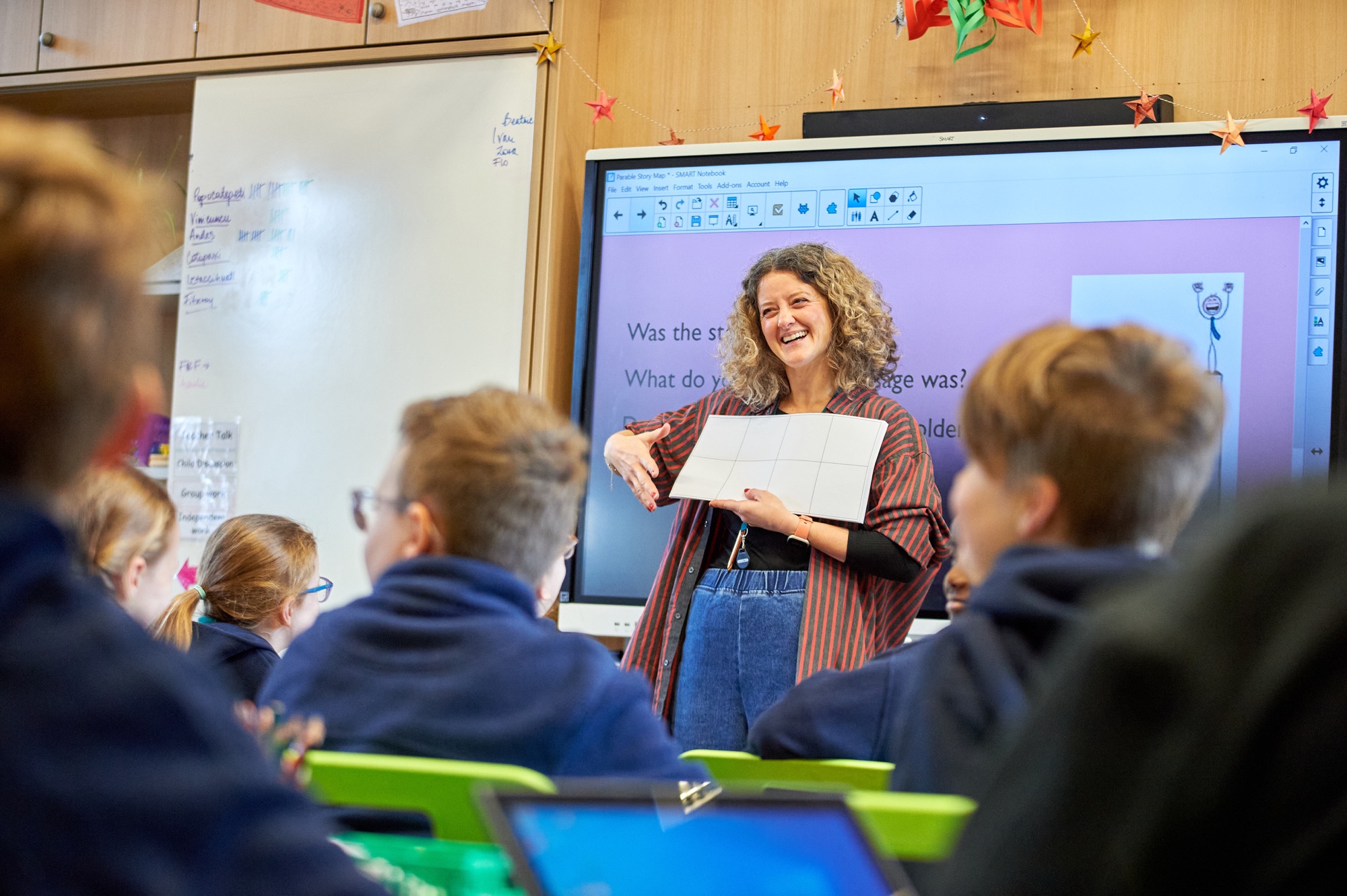 Teacher smiling at the front of a classroom, holding up a worksheet to her pupils