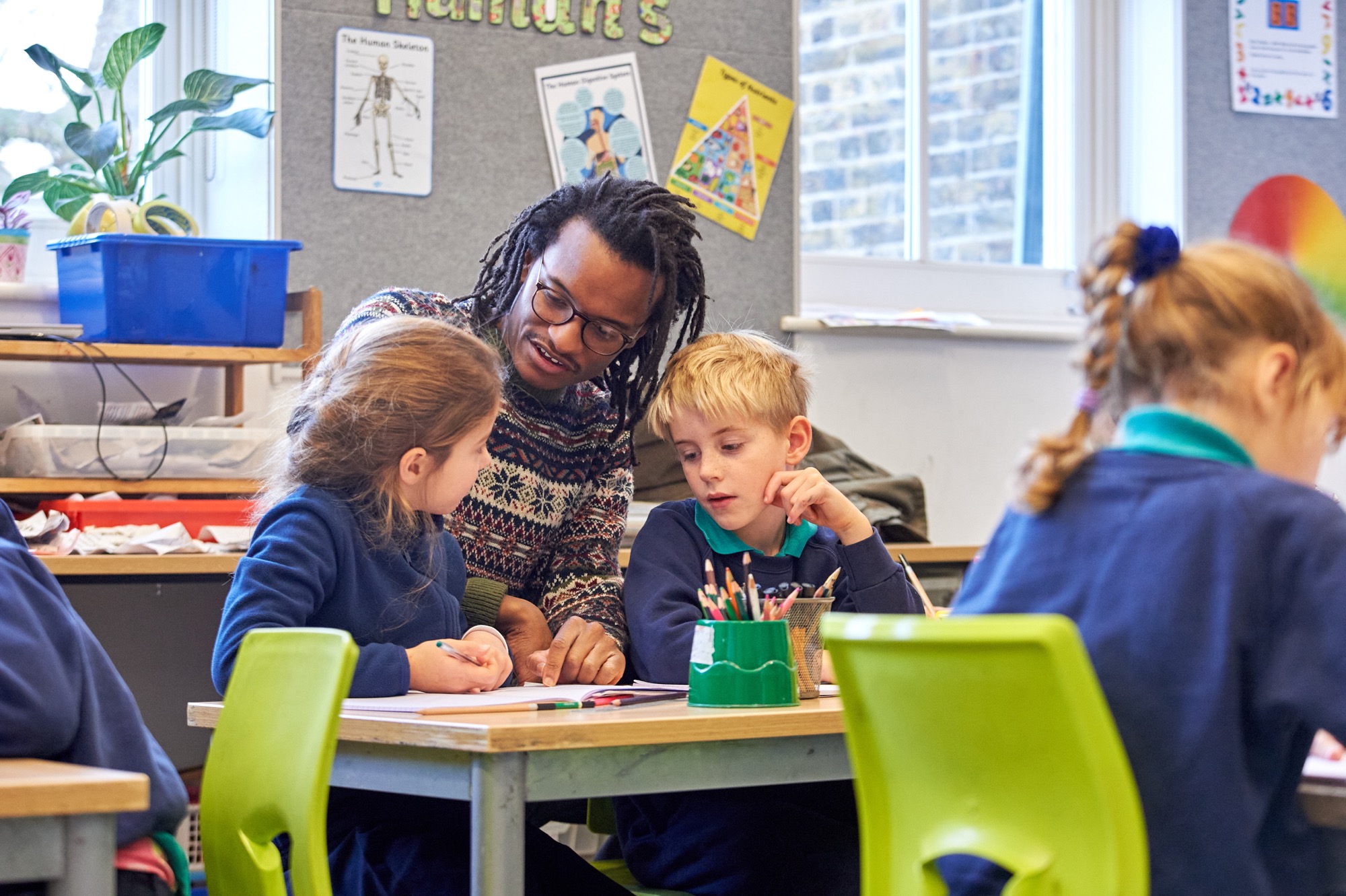Classroom adult supporting two primary pupils with their work at a shared table