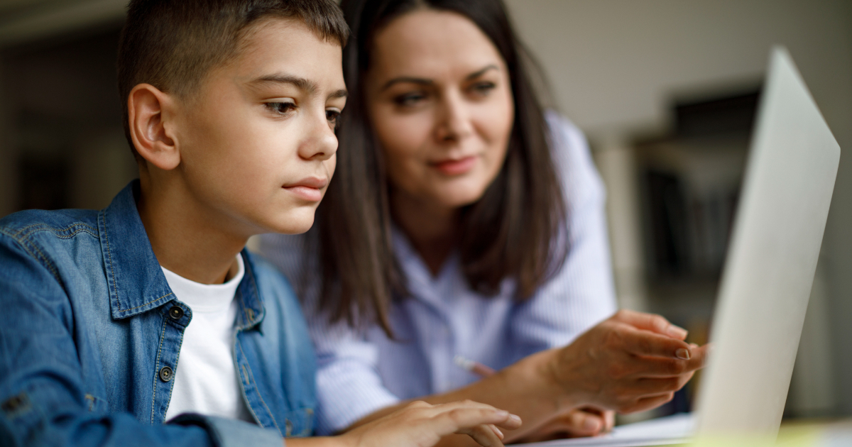 A young student works at a laptop while a teacher points at the screen