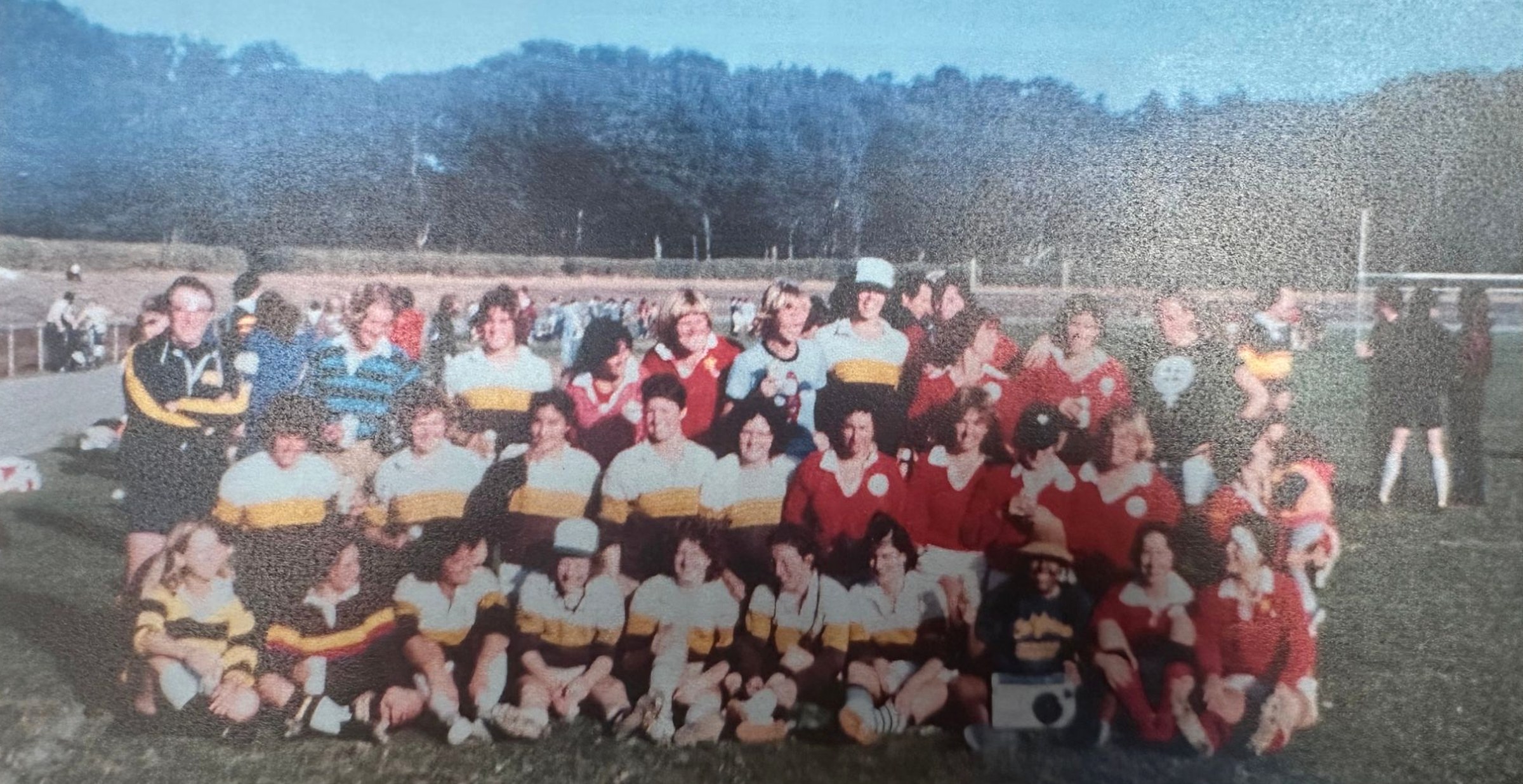 A vintage team photo of the Seattle Seabyrds Women's Rugby Football Club and their opponents, gathered together on a rugby pitch after a match, in hooped and red jerseys
