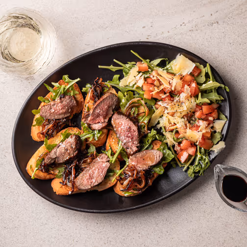 Black plate with slices of seared lamb on toasted bread with arugula and caramelised onions, served with a side salad of chopped tomatoes, arugula, and shaved cheese, accompanied by a glass of white wine and a small container of dark sauce.