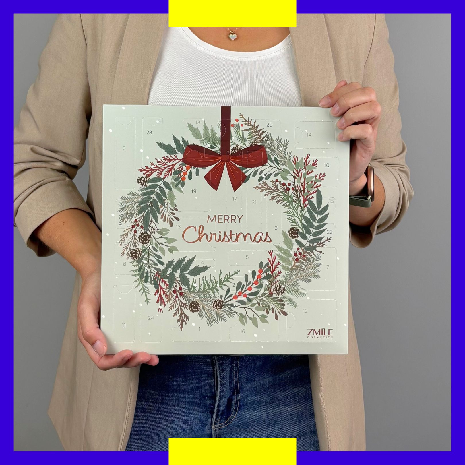 Person zeigt das Größenverhältnis des quadratischen Beauty Adventskalenders Jingle and Glow von ZMILE Cosmetics mit Weihnachtskranz vor grauem Hintergrund.