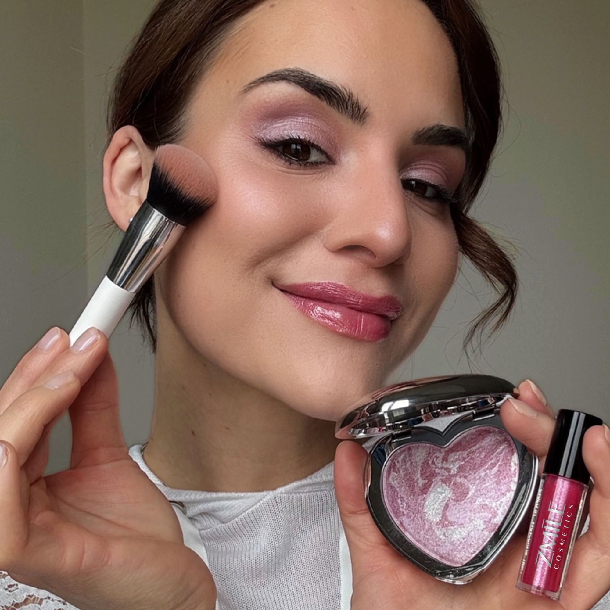 Junge Frau in Nahaufnahme hält Lipgloss und Blush in der einen Hand - Kosmetikpinsel in Anwendung am hohen Wangenknochen