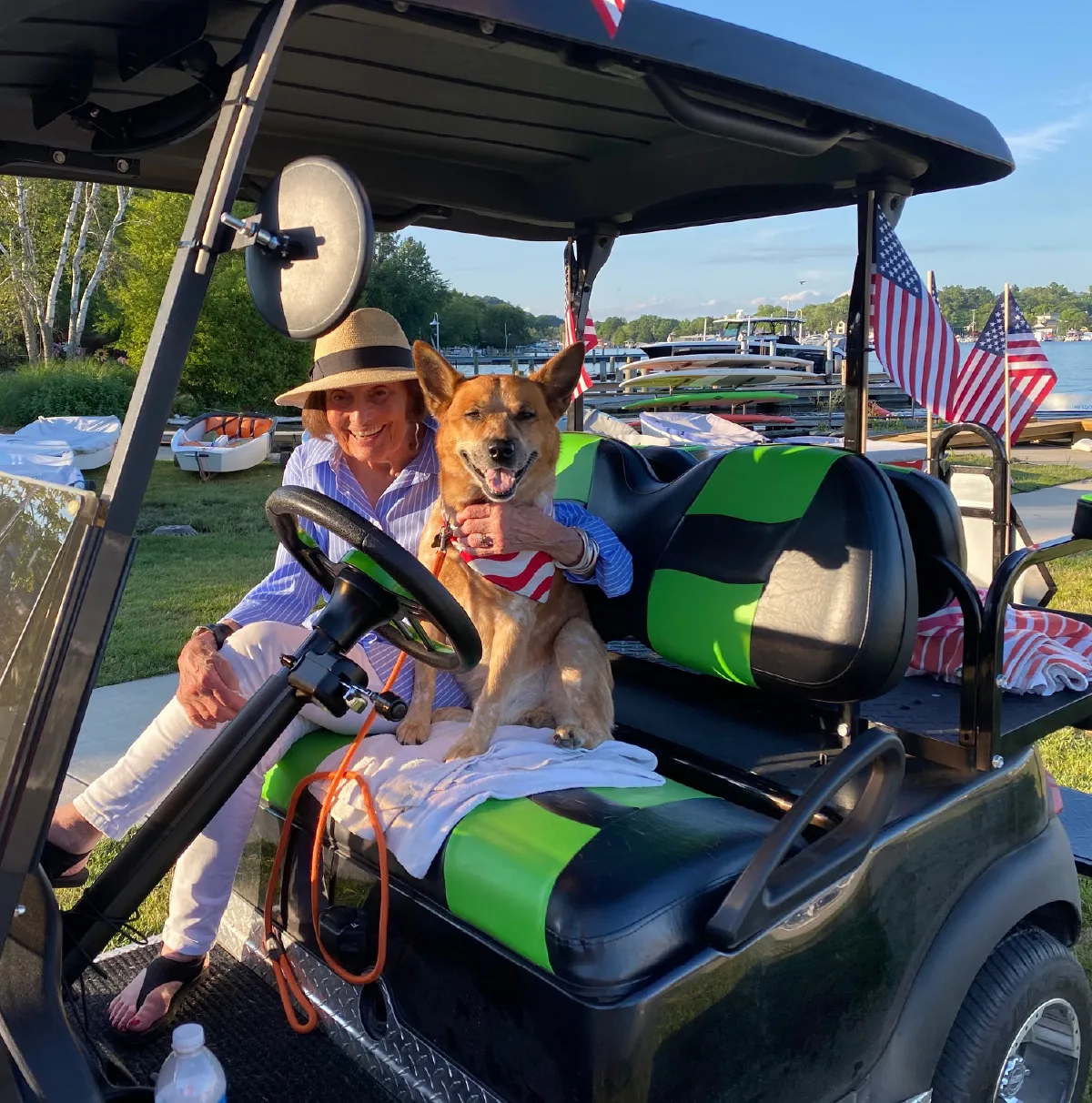 Smiling woman in a hat sitting beside a dog wearing a patriotic bandana on a green and black golf cart near a lakeside with boats and American flags.