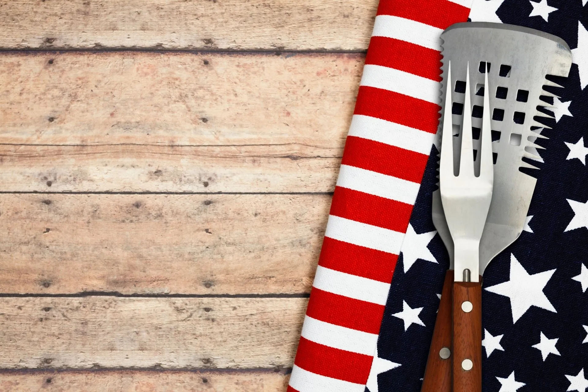 BBQ fork and spatula with wooden handles resting on an American flag-themed cloth on a wooden surface.