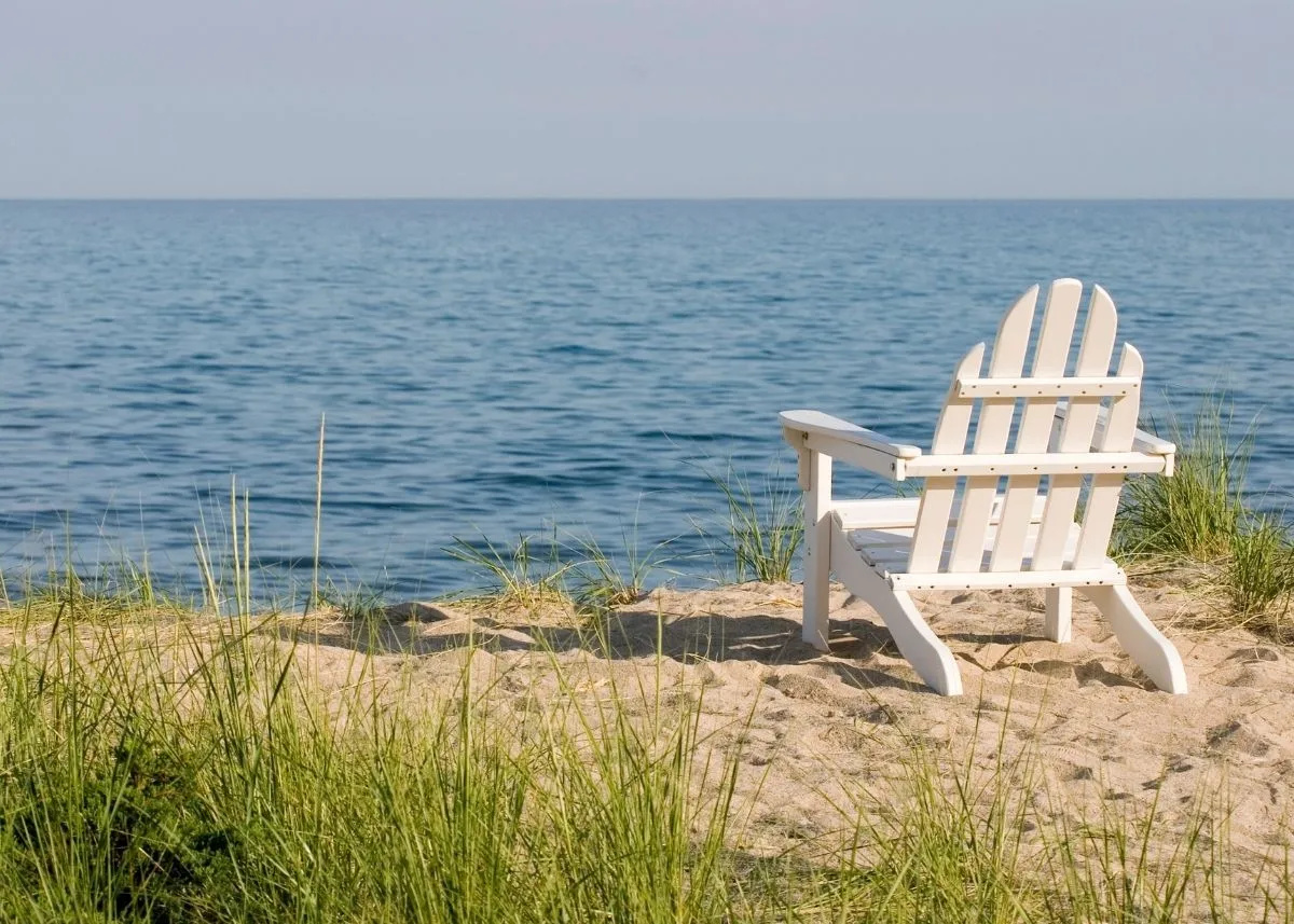 White Adirondack chair on sandy beach facing calm blue lake under clear sky.