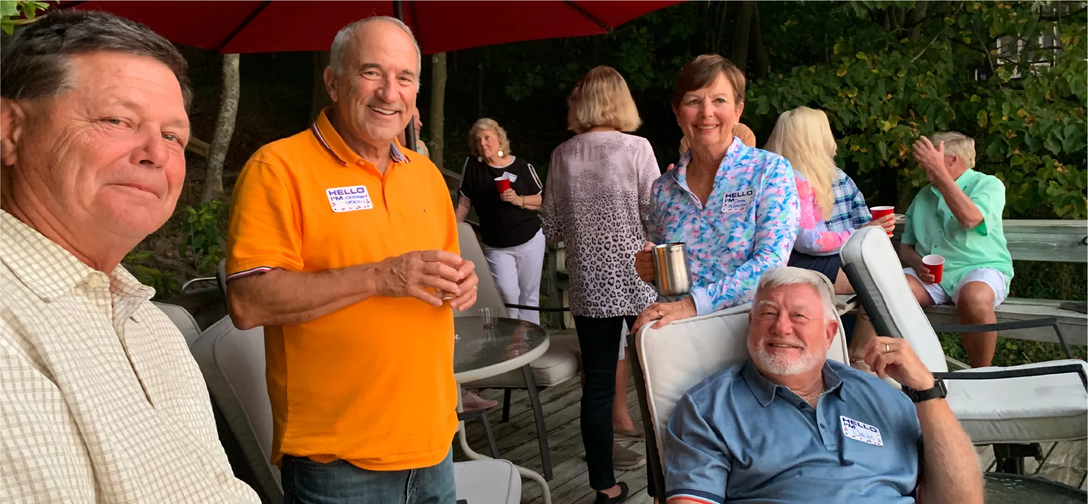 Group of smiling older adults socializing outdoors on a wooden deck with drinks, some wearing name tags, under a red umbrella.