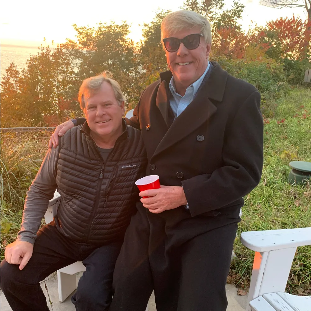 Two middle-aged men smiling outdoors at sunset, one wearing sunglasses and holding a red cup with fall foliage in the background.