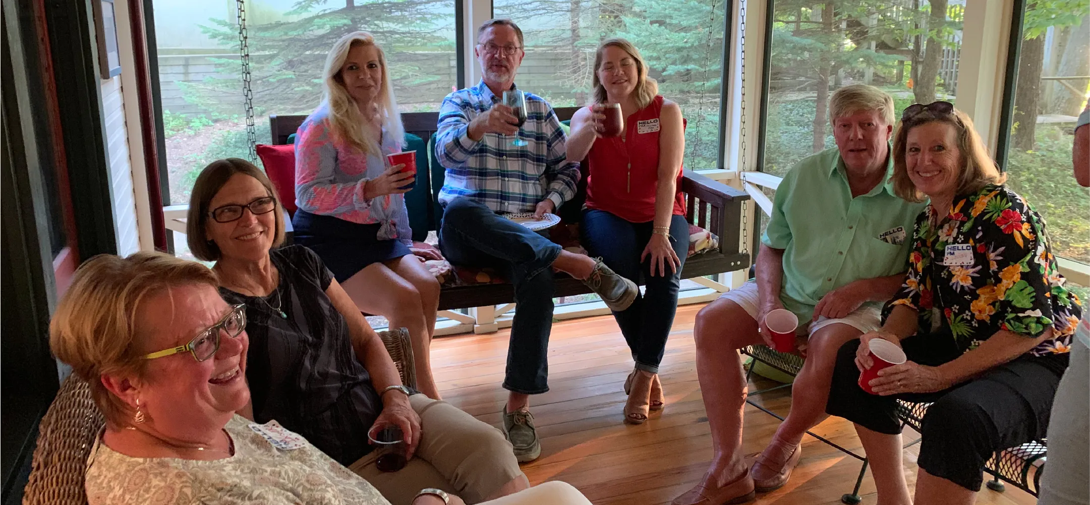 Group of seven adults sitting and smiling on a wooden porch with drinks, surrounded by trees outside.